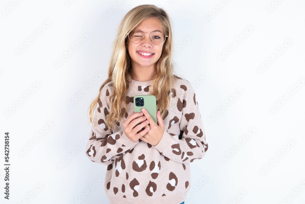 Pleased beautiful caucasian teen girl wearing animal print sweater over white wall using self phone and looking and winking at the camera. Flirt and coquettish concept. Pleased beautiful caucasian teen girl wearing animal print sweater over white wall using self phone and looking and winking at the camera. Flirt and coquettish concept.