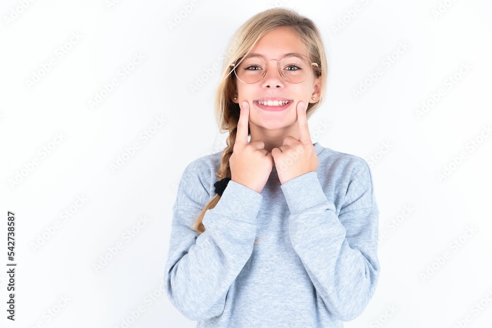 Happy beautiful caucasian teen girl wearing gray sweater over white wall with toothy smile, keeps index fingers near mouth, fingers pointing and forcing cheerful smile Happy beautiful caucasian teen girl wearing gray sweater over white wall with toothy smile, keeps index fingers near mouth, fingers pointing and forcing cheerful smile
