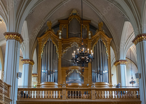 Finland, Kotka - July 18, 2022: Kotka-Kymin Parish Church or Seurakuntayhtymä. The entire gold and silver organ in closeup under Gorhic-revival ceiling and pillars. Hanging light fixture