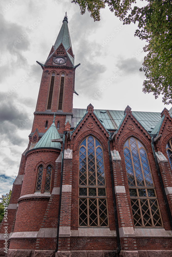 Finland, Kotka - July 18, 2022: Kotka-Kymin Parish Church or Seurakuntayhtymä. North red brick facade with clock tower against gray cloudscape. Gothic-revival windows and green roof