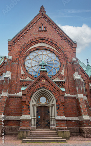 Finland, Kotka - July 18, 2022: Kotka-Kymin Parish Church or Seurakuntayhtymä. Closeup of North side entrance block red brick facade with rose window and green cross under blue sky photo