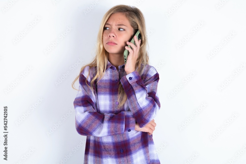 Sad beautiful caucasian teen girl wearing plaid purple shirt over white wall talking on smartphone. Communication concept. Sad beautiful caucasian teen girl wearing plaid purple shirt over white wall talking on smartphone. Communication concept.