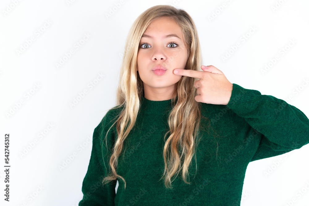 Charming beautiful caucasian teen girl wearing green knitted sweater over white wall pointing on pout lips with forefinger, showing effect after lifting procedure,