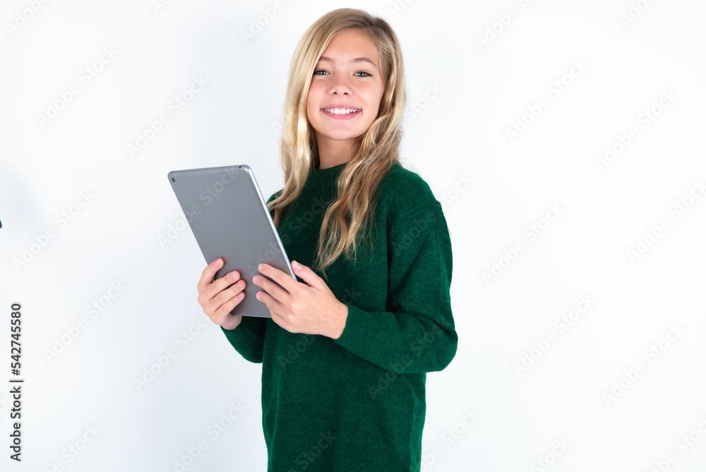 Photo of optimistic beautiful caucasian teen girl wearing green knitted sweater over white wall hold tablet Photo of optimistic beautiful caucasian teen girl wearing green knitted sweater over white wall hold tablet