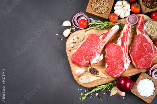 Many raw ribeye beef steaks ready for grilling with seasonings and vegetables on wooden cutting board over dark concrete background. Cowboy steak ready for grilling. Top view