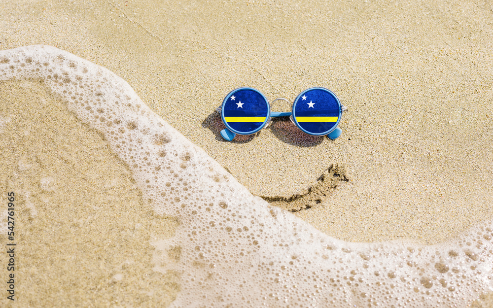 Obraz premium Sunglasses with flag of Curacao on a sandy beach. Nearby is a sea lightning and a painted smile. The concept of a successful vacation in the resorts of the Curacao.