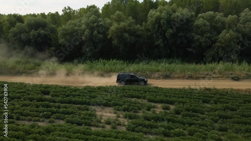 Aerial view of an 4x4 SUV driving on a dusty road in the countryside. Footage was shot from a drone at a lower altitude flying on a side of the sport utility vehicle keeping it in view.