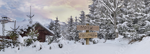 merry christmas written on wooden postsign  next to an alpine chalet and forest in snow photo