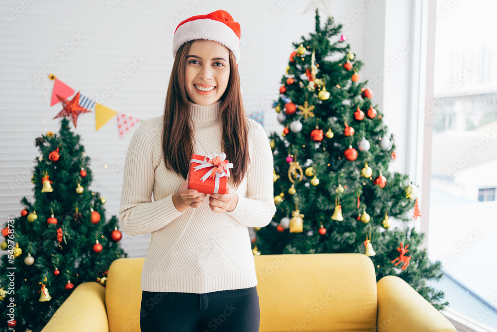 Young Asian woman wearing a sweater and Santa Claus hat smiles and holds a Christmas gift in a red box with ribbon in her hands a living room with a decorated Christmas tree in the background. Young Asian woman wearing a sweater and Santa Claus hat smiles and holds a Christmas gift in a red box with ribbon in her hands a living room with a decorated Christmas tree in the background.