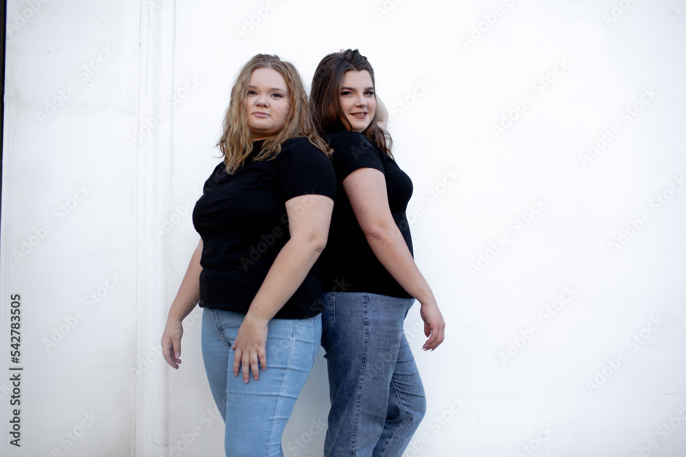 Two beautiful plump women wearing jeans and t-shirt, friends posing at the background against white street wall Two beautiful plump women wearing jeans and t-shirt, friends posing at the background against white street wall