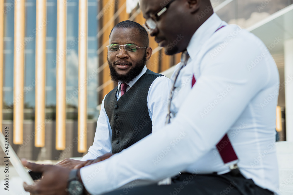 Business meeting outdoors. Two dark-skinned men in suits sit on the steps of a city building with a laptop and have a conversation. Friendly support and brainstorming. Partnerships Business meeting outdoors. Two dark-skinned men in suits sit on the steps of a city building with a laptop and have a conversation. Friendly support and brainstorming. Partnerships