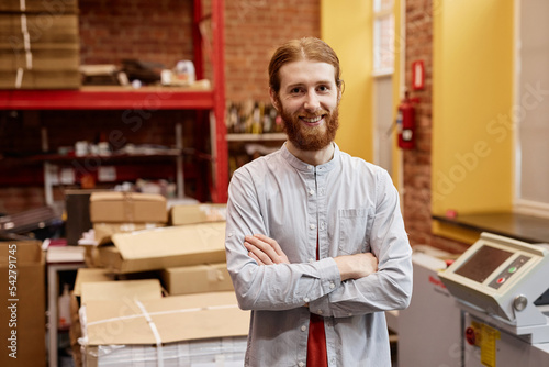 Waist up portrait of smiling young man working in industrial printing shop and standing with arms crossed, copy space