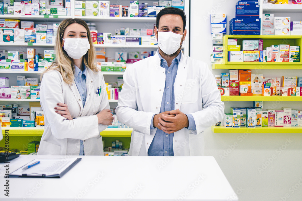 Male and female pharmacy workers on duty, wearing masks and sorting medicaments on shells.
Talking to each other and writing down names of medical drugs