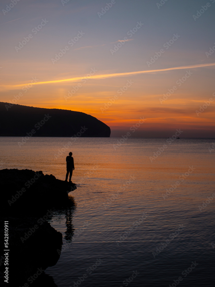 Young male watching sunrise over the sea from a rocky beach. Moraira, Calpe, Costa Brava.