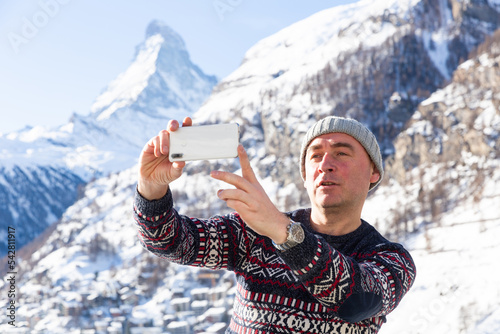 Adult man in warm knitted sweater and hat traveling in winter Swiss Alps, recording video with mobile phone outdoor on sunny day photo