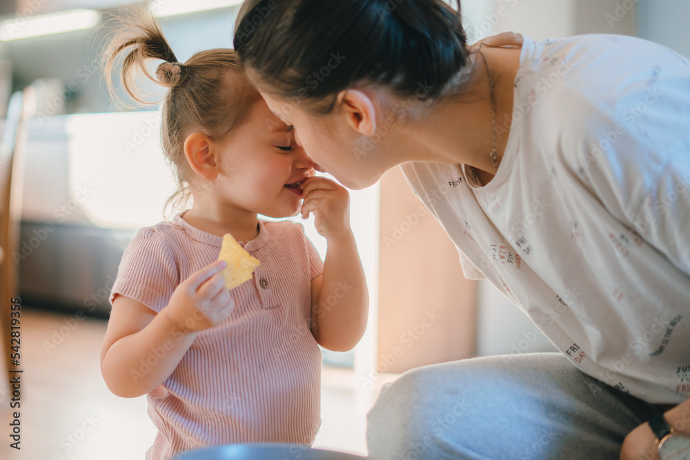 Caring Caucasian mother kissing her little girl on the nose while the little girl eats a cookie. Cute little girl with a cookie in her mouth. Children's Caring Caucasian mother kissing her little girl on the nose while the little girl eats a cookie. Cute little girl with a cookie in her mouth. Children's