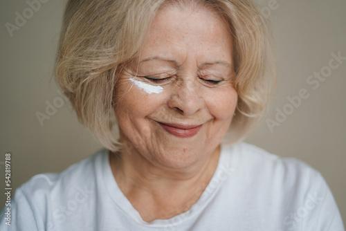 Head shot of beautiful smiling senior woman putting on the creme. Wrinkle free with natural anti-aging cosmetics. Product for beauty skincare, makeup