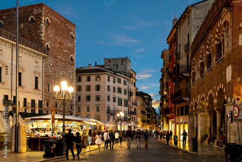 Fototapeta Naklejka Na Ścianę i Meble -  Tourists and Italians visit the outdoor market, shops and cafes at the illuminated Piazza delle Erbe, in the historic center of Verona, Italy.