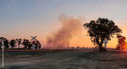Panoramic image of a plume of balck smoke rising at sunset from a line of wild fire in the distant farmland, railway crossing and gravel road, rural Victoria, Australia