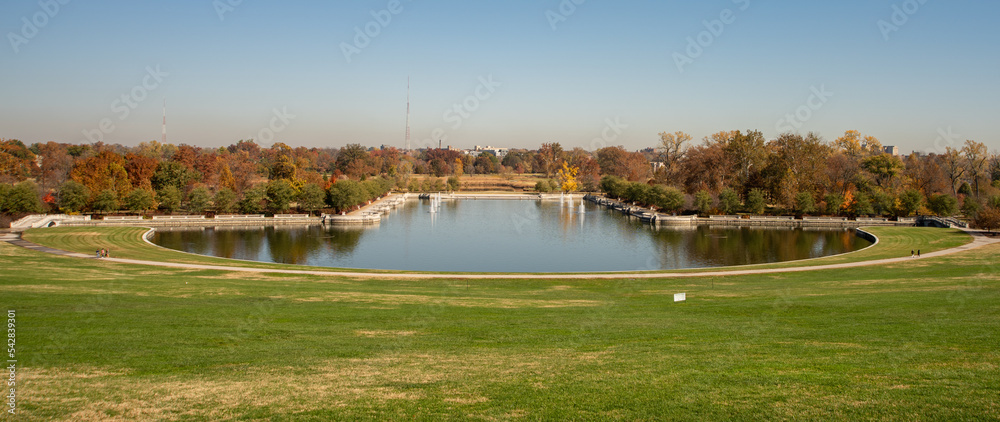 Large lake with pedestrian walking path around it and water fountains. A large expanse of grass is in front and bridges are on either side. Fall trees with leaves of red, green, orange and yellow.