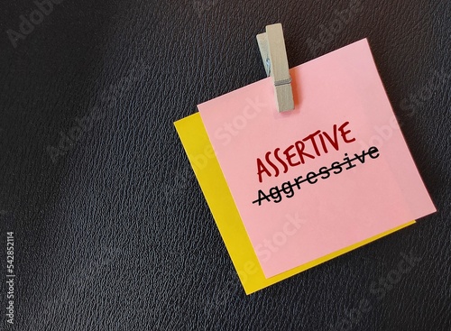 Pink note on black copy space background with text written AGGRESSIVE crossed off to ASSERTIVE, means to develop assertiveness or ability to express opinion positively with honest confidence  photo