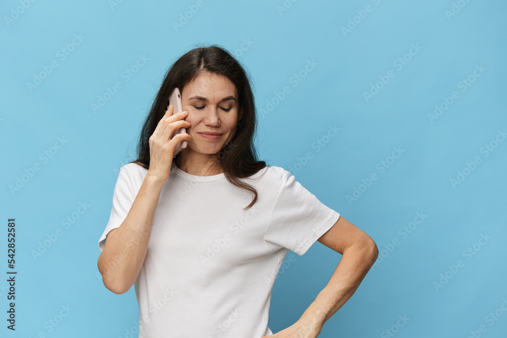 portrait of a beautiful, pleasant woman with long hair, in a light t-shirt holding a fashionable smartphone in her hands during a call, closing her eyes. Horizontal photo on a pink background portrait of a beautiful, pleasant woman with long hair, in a light t-shirt holding a fashionable smartphone in her hands during a call, closing her eyes. Horizontal photo on a pink background