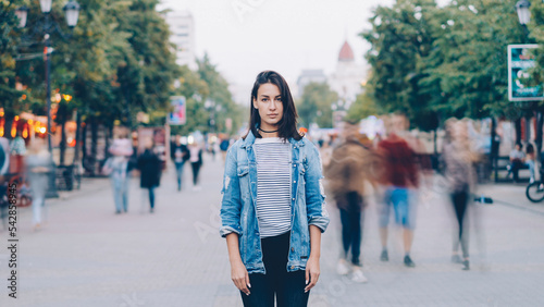portrait of tired young woman student standing alone in city center and looking at camera with straight face while crowds of men and women are whizzing around.