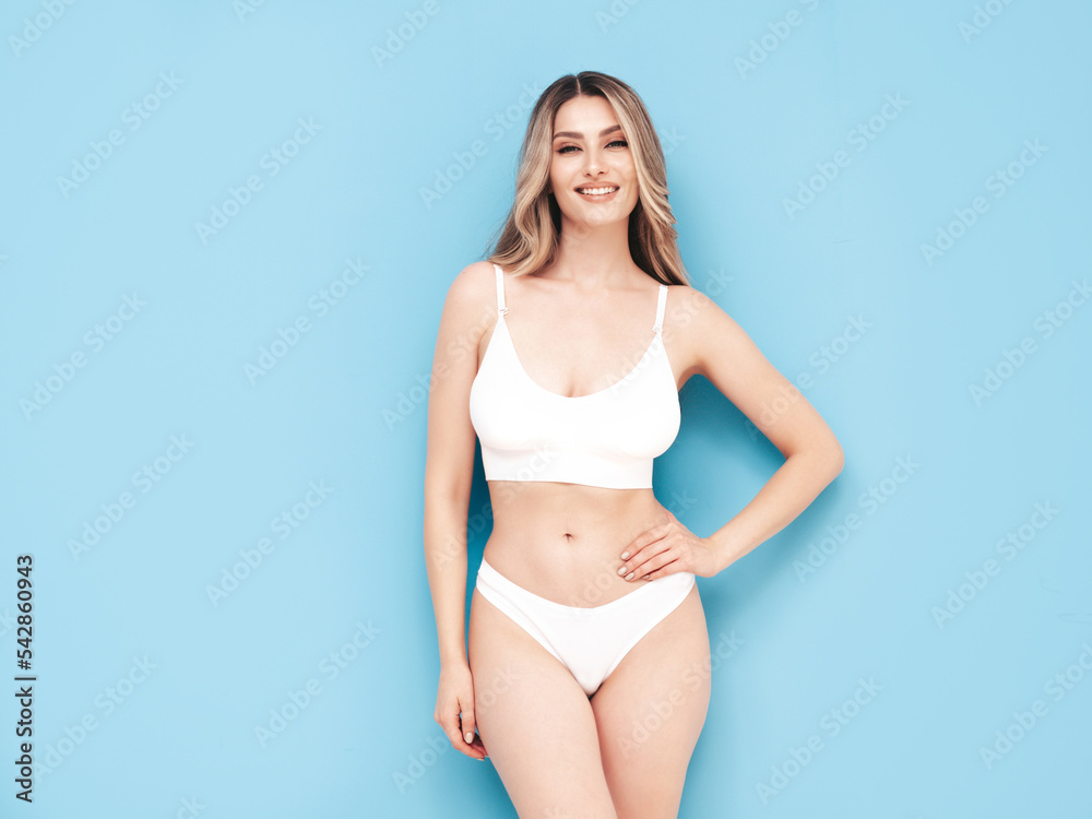 Fashion portrait of young beautiful woman. Attractive carefree blond model wearing pure white lingerie. Hot tanned smiling blonde posing near blue wall in studio, showing sexy natural body Fashion portrait of young beautiful woman. Attractive carefree blond model wearing pure white lingerie. Hot tanned smiling blonde posing near blue wall in studio, showing sexy natural body