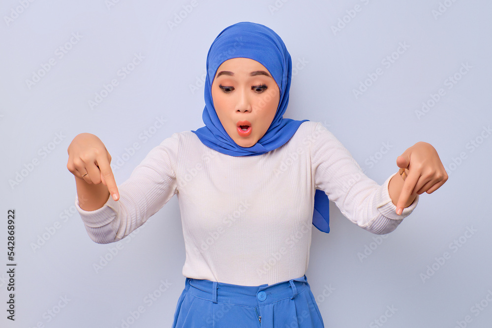 Excited young beautiful Asian Muslim woman pointing fingers down, inviting people to follow isolated over white background Excited young beautiful Asian Muslim woman pointing fingers down, inviting people to follow isolated over white background