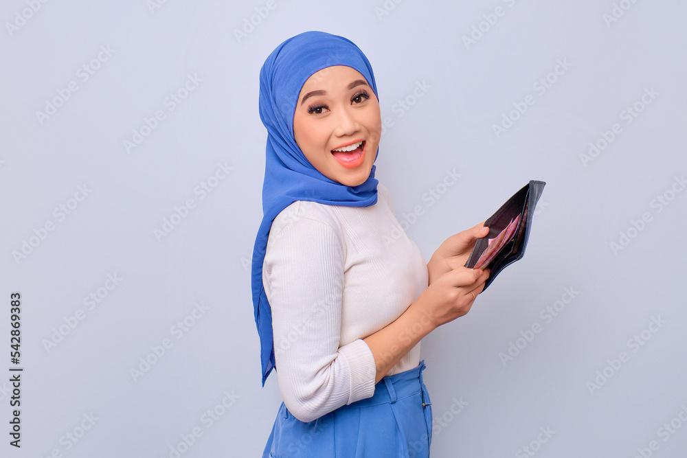 Excited young beautiful Asian Muslim woman showing wallet full of money banknotes isolated over white background Excited young beautiful Asian Muslim woman showing wallet full of money banknotes isolated over white background