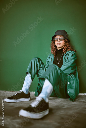 Fashion, style and clothes with a model black woman sitting on the floor against a green background. Portrait, glasses and fashionable with a young female posing to promote trendy or edgy clothing