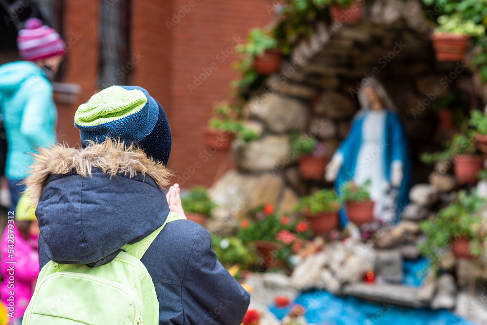 a boy stands in front of a statue of the Holy Virgin Mary and prays	