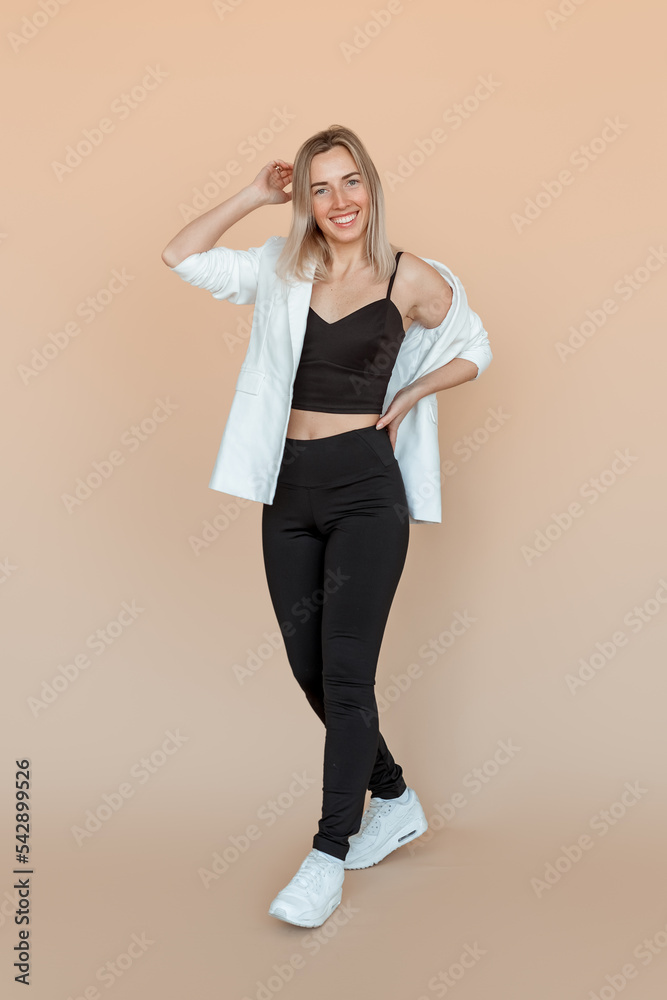 Full length of smiling young woman with freckles posing in white blazer and black clothes with carefree upbeat face expression. Indoor studio shot on beige background Full length of smiling young woman with freckles posing in white blazer and black clothes with carefree upbeat face expression. Indoor studio shot on beige background