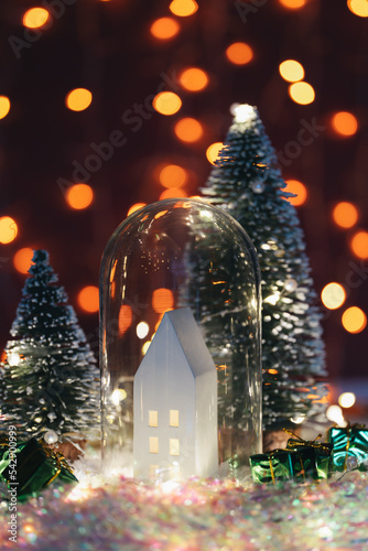 Christmas cute little house in glass dome with tree in snow. Glowing festive garland on dark background. Cozy atmosphere with home decor with selective focus. New Year card © Alex Shi