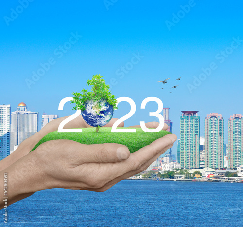 2023 white text with planet and tree on green grass field in hands over city tower, Happy new year 2023 ecological cover, Save the earth concept, Elements of this image furnished by NASA