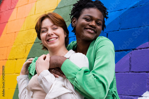 Happy woman embracing girlfriend in front of rainbow flag painted on wall photo