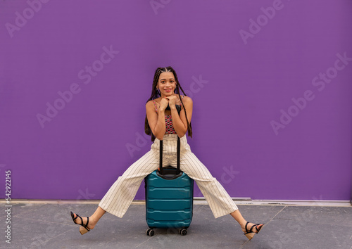 Smiling young woman sitting on suitcase in front of purple wall photo