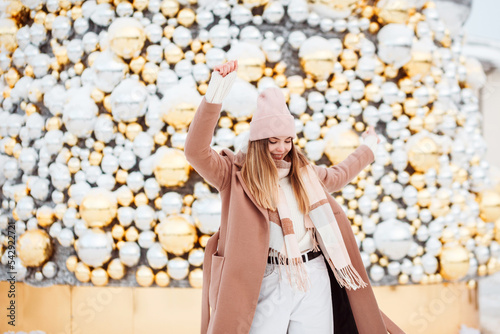 Cute girl in stylish winter beige outfit having fun outdoors in front of christmas tree decorated with silver and golden balls. 
