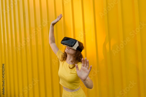 Smiling woman wearing virtual reality simulator gesturing in front of yellow wall photo