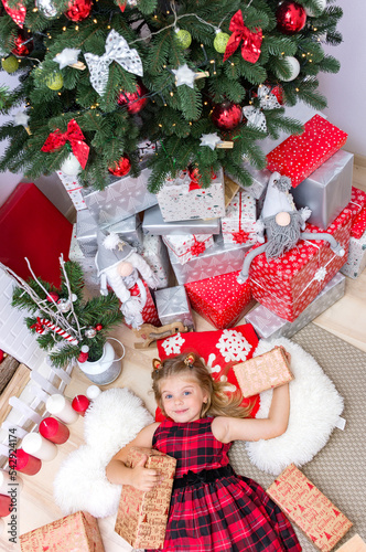 Happy little smiling girl with a Christmas gift box is lying under the Christmas tree