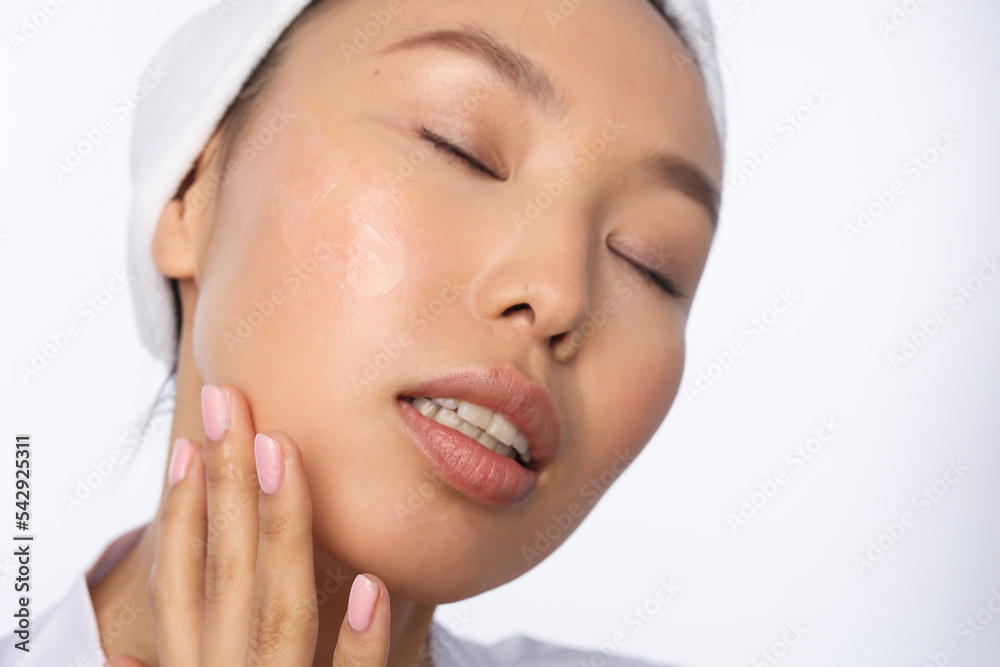 Closeup of asian woman with towel on head apply white cream on face. Young girlportrait. Neutral natural pink manicure on nails. Woman doing cosmetic procedure, white background, isolated. Closeup of asian woman with towel on head apply white cream on face. Young girlportrait. Neutral natural pink manicure on nails. Woman doing cosmetic procedure, white background, isolated.