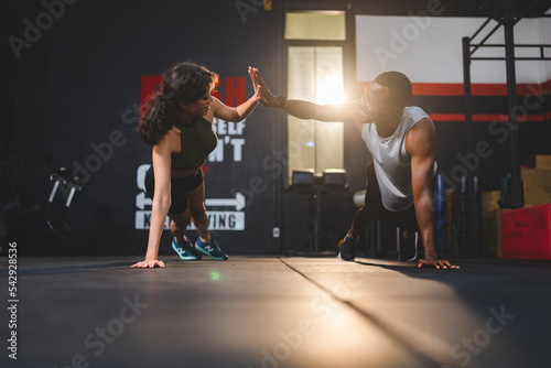 Young fit and health conscious girlfriend and boyfriend clapping hands while practicing single hand balance planks pose in modern dark gym floor in sportswear while training together © chokniti