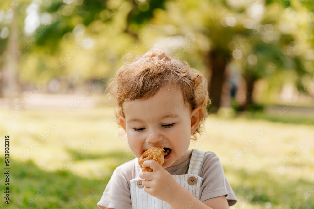 Close up outdoor photo of little cute curly girl eating croissant in the park in summer sunny day. Cute little girl in red attire running in park. Charming kid with curly hair playing at lawn. Close up outdoor photo of little cute curly girl eating croissant in the park in summer sunny day. Cute little girl in red attire running in park. Charming kid with curly hair playing at lawn.