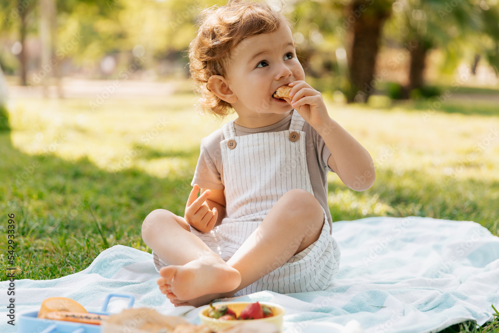 Outdoor portrait of charing cute little girl with curls sitting in the park inn sunny warm day and eating croissant. happy kid in summer suit sitting on the grass and having lunch