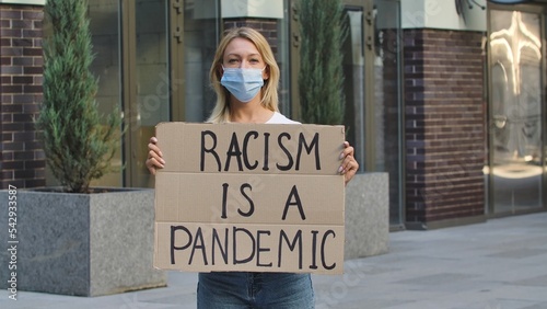Young woman in medical mask stands with a cardboard poster RACISM IS A PANDEMIC in a public place outdoor. No racism but tolerance and political correctness in society.