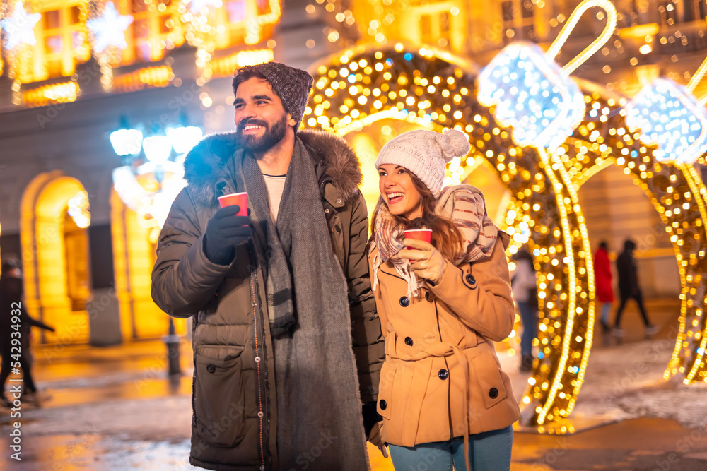 Obraz premium Couple drinking mulled wine and walking down decorated city streets while celebrating Christmas