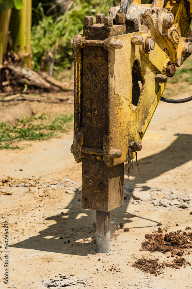 An excavator fitted with a hydraulic breaker punching holes into a portion of a damaged street, breaking it apart. Road demolition in progress.