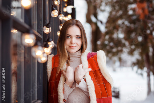 Romantic portrait of beautiful young woman in winter time. Girl in warm woolen pachwork vest standing next to window with garland of lights. Christmas holiday time.