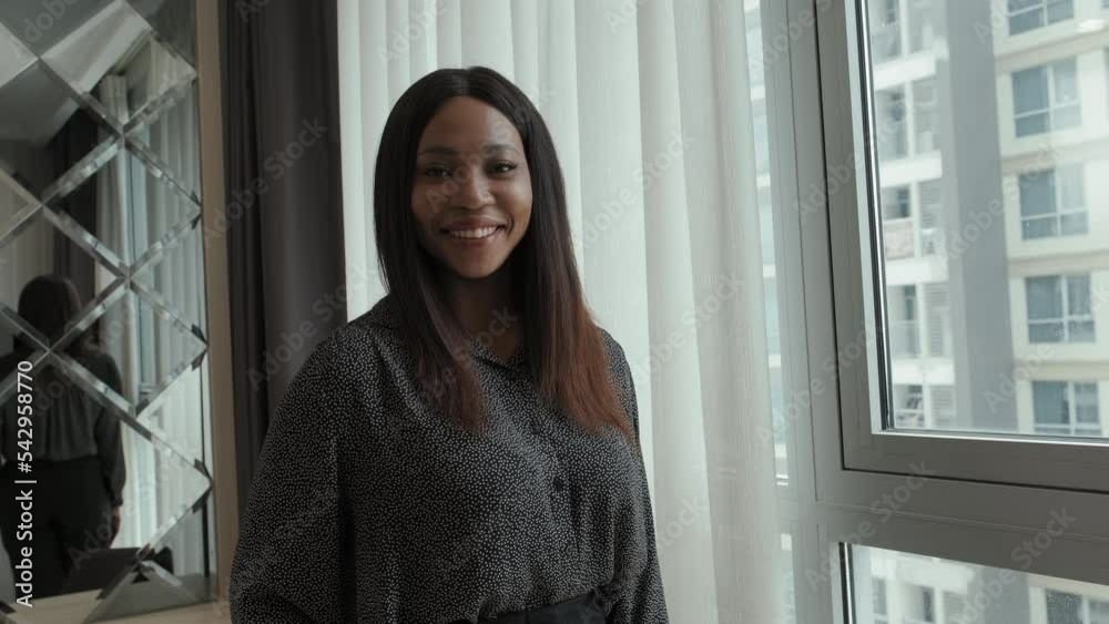 Medium slowmo portrait of attractive young African American businesswoman smiling at camera standing by window in contemporary hotel room Medium slowmo portrait of attractive young African American businesswoman smiling at camera standing by window in contemporary hotel room