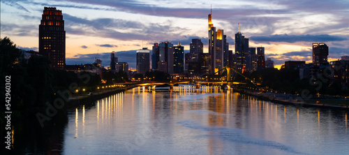 Frankfurt wide angle nighttime panorama. German city with downtown skyline, tall buildings  and bridge. Evening sky with clouds and reflection in River Main at blue hour with colorful illumination.